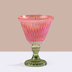 A ribbed glass candle holder with a petal pink ribbed glass and orange iridescent upper bowl and a green pedestal base, shown on a white surface against a beige background.