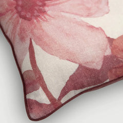 A close-up of a square throw pillow featuring a watercolor-style floral print in shades of dusty pink and burgundy on a cream background, finished with a dark burgundy piped edge.