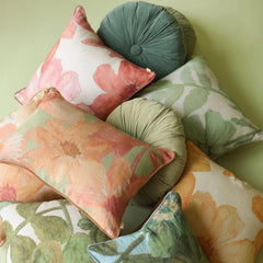 A styled collection of decorative throw pillows with floral and botanical patterns in soft sage green, dusty teal, coral pink, and peach tones, arranged against a solid pale green background.