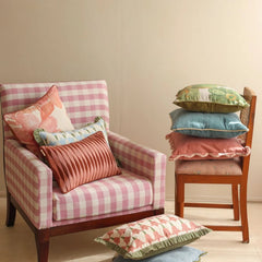A pink and white checkered armchair with decorative throw pillows, alongside a wooden chair stacked with additional patterned cushions, set against a plain beige wall.