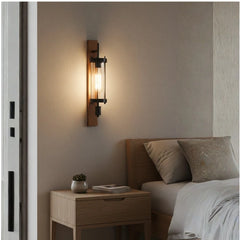 A modern wall-mounted light fixture with a vertical wooden backplate, clear glass shade, and exposed Edison bulb, shown lit in a bedroom setting next to a bed and nightstand.