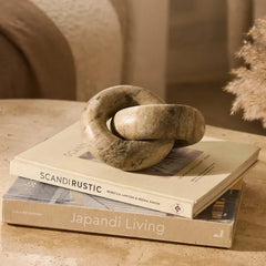 Moro Chain - a round, knot-like stone sculpture in a natural beige and brown textured finish, displayed on top of two stacked books in a warm, rustic interior setting. 