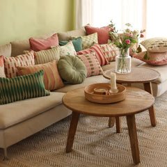 A styled living room scene with a beige sectional sofa covered in multiple patterned throw pillows in green, pink, red, beige, and a Cuddle Fern Velvet Round Cushion.