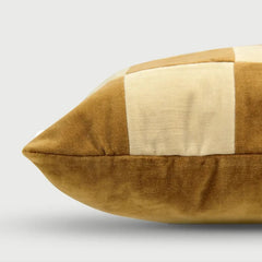 A close-up of a square throw pillow with a patchwork design, featuring a mustard yellow velvet fabric and a cream-colored linen panel, set against a light gray background.