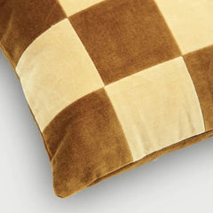 A close-up of a square throw pillow featuring a checkered pattern in warm brown and cream velvet fabric, set against a neutral light gray background.