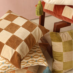 A styled arrangement of decorative throw pillows in a warm neutral setting. The most prominent pillow features a large-scale checkered pattern in brown and cream velvet, while other pillows in the scene display patchwork designs in olive green, cream, and rust red, with one pillow showing a tan and blue geometric pattern.