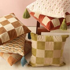 A styled collection of decorative throw pillows in warm neutral and earthy tones, including checkered and patterned designs with tassels, arranged on a chair and floor against a soft pink wall.