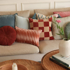 A styled collection of decorative throw pillows arranged on a beige sofa. The pillows include a round deep red velvet pillow, a red and cream striped pillow, a red and cream checkered pillow, a blue velvet pillow, a light sage green pillow, and a striped beige and blue pillow.