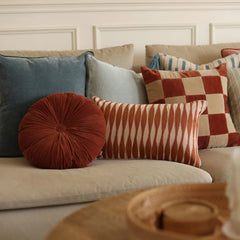 A styled collection of home decor throw pillows arranged on a light beige sofa in a living room setting. The pillows include a round rust-red velvet pillow, a striped rust-and-cream pillow, a checkered red and beige pillow, and a slate blue pillow.