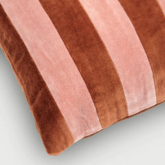 A close-up of a striped velvet throw pillow in alternating shades of medium brown and dusty pink, shown against a light gray background.