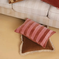 A pink striped velvet throw pillow resting on a brown fringed cushion on a beige floor, with a cream sofa in the background.