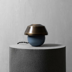 A modern table lamp with a matte blue spherical base and a brushed bronze dome-shaped shade, displayed on a textured gray cylindrical pedestal against a light gray wall.