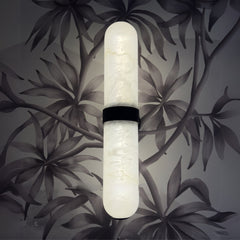 A wall-mounted light fixture with a white alabaster tube and a black band, set against a floral patterned backdrop.