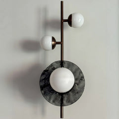 A wall sconce featuring a vertical metal rod with two white opaline glass globes on either side, and a larger slumped glass circle in the center.