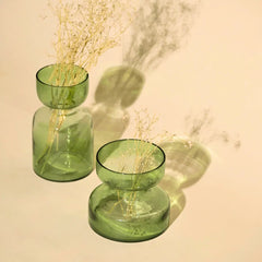 Two translucent sage green glass vase-like candle holders with a flared rim, each containing dried pale stems, arranged on a warm beige background with soft shadows.