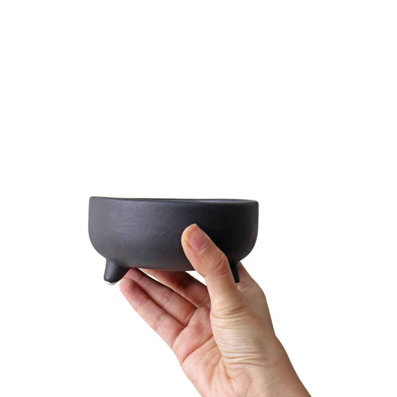 A hand holding a black ceramic Pluto Bowl with three tiny legs on a plain background.