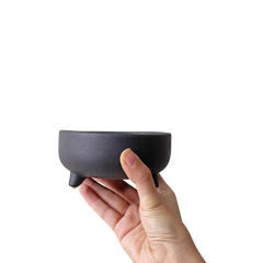 A hand holding a black ceramic Pluto Bowl with three tiny legs on a plain background.