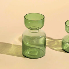 A sage green glass vase with a flared rim and a rounded cylindrical body, shown on a warm beige background with soft shadows.