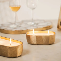 Fleur Votive, lit candles in gold wooden holders on a table with blurred wine glasses in the background.