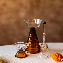 A set of three colored glass items, including a tall diya, a short diya, and a ringing bell, placed on a table with a textured background. The items appear to be made of borosilicate glass with a colored finish.
