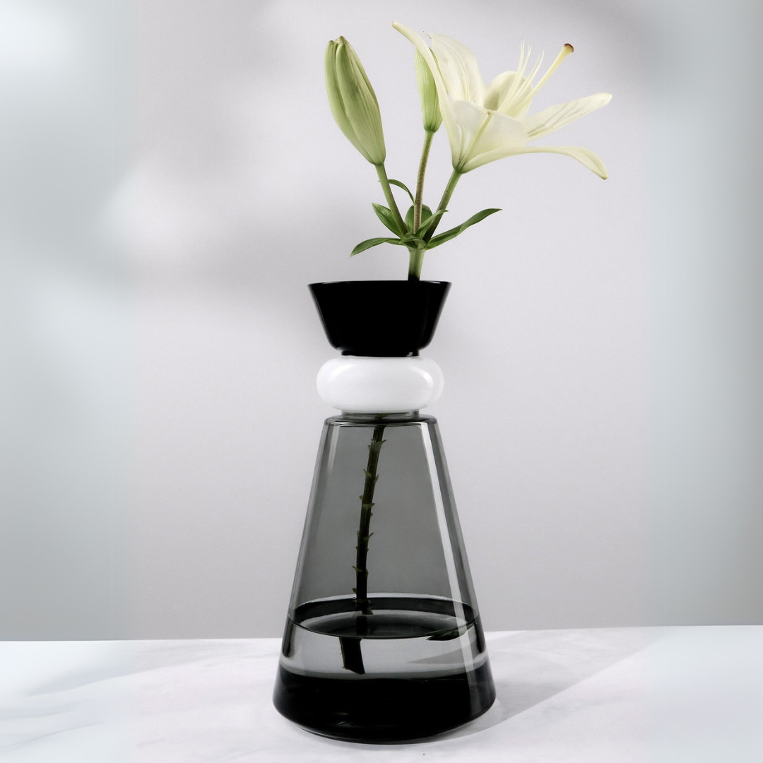 Clear glass vase with black base and white top, containing a plant with white flowers on a light gray background.
