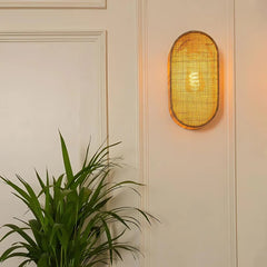 Wall-mounted light fixture with woven design on a neutral wall with a plant in the foreground.