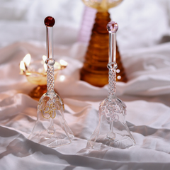 Two ornate glass bells on a soft white fabric background with a blurred candle and bottle in the background.