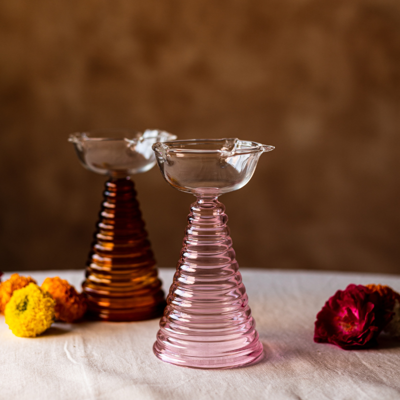 Two tall glass diya holders in amber and pink colors with a textured design, displayed on a table with a few flowers scattered around.