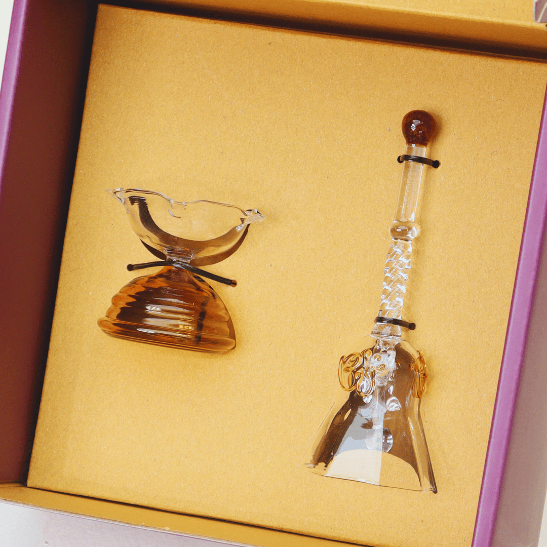 A set of two glass items, a ringing bell and a short diya, packaged in a gift box with a yellow and purple interior.