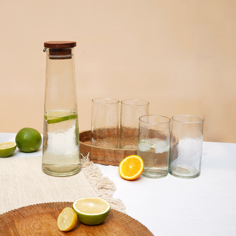 A set of clear glassware with a hammered texture, displayed alongside a carafe filled with liquid, and citrus fruits on a wooden surface.