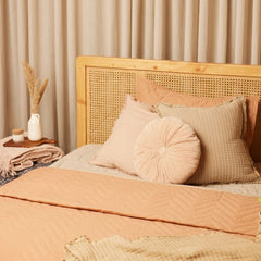 A cozy bed styled with peach and beige bedding, including a quilted peach blanket, textured beige sheets, a round pleated blush pink pillow, and a beige checkered pillow, set against a light wood cane headboard with beige curtains in the background.