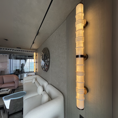 A alabaster wall sconce with a cylindrical marble tube and LED lighting, mounted on a wall in a room with a light-colored sofa.