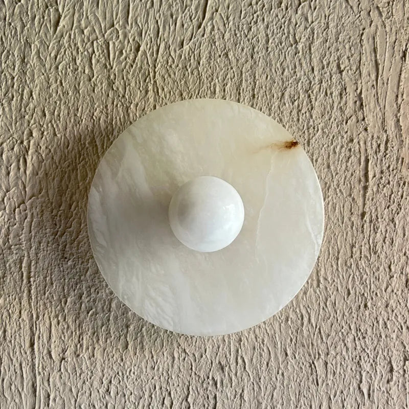 A round white alabaster stone or resin wall hook with a central spherical protrusion, mounted on a textured beige plaster wall.
