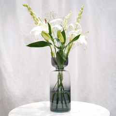 A tinted glass vase with a retro-inspired shape, featuring a gradient of vibrant colors, filled with water and a bouquet of white flowers.