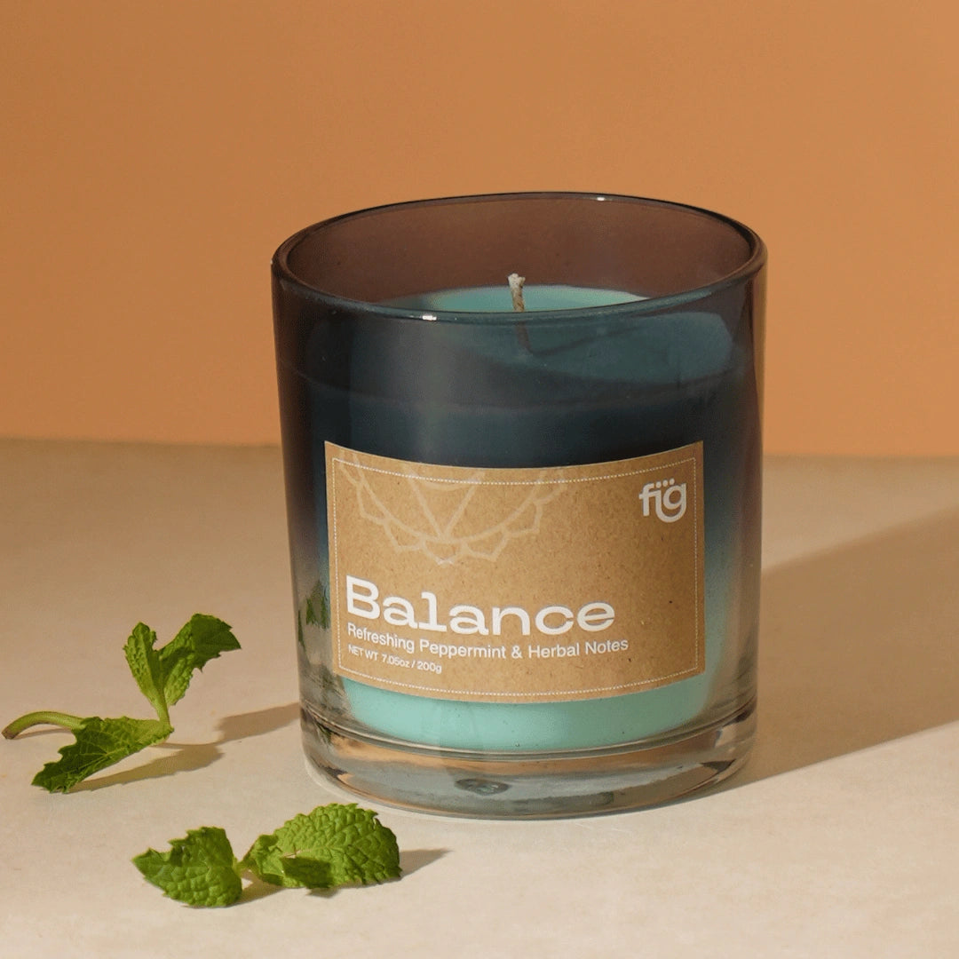 A clear glass candle with Balance Peppermint Vegan Wax Candle, labeled 'BALANCE'. A small sprig of mint is also visible to the side.
