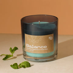 A clear glass candle with Balance Peppermint Vegan Wax Candle, labeled 'BALANCE'. A small sprig of mint is also visible to the side.