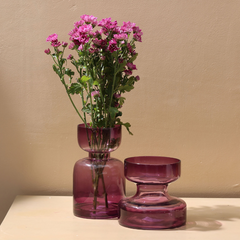 A tall lilac colored glass vase with a retro shape, displayed with a smaller vase of the same color in the background, both containing pink flowers.