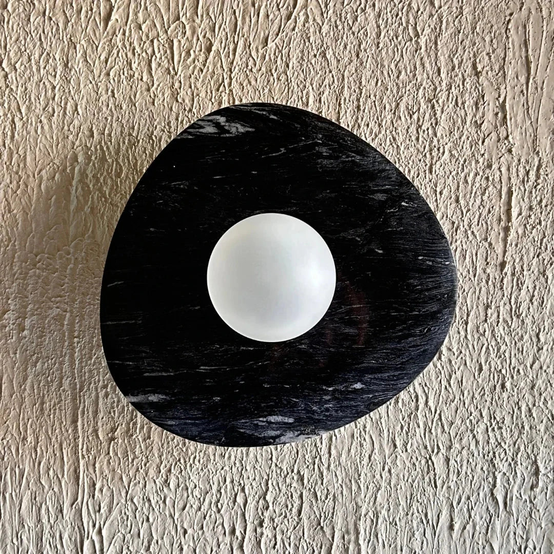 A round textured black wall light with a matte finish, hanging on a wall with a white wall texture in the background.