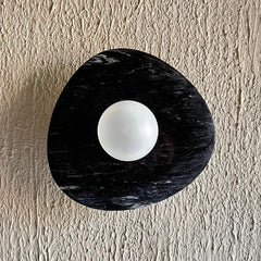 A round textured black wall light with a matte finish, hanging on a wall with a white wall texture in the background.