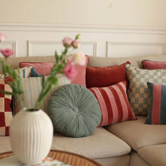A cozy living room scene with a beige sofa styled with multiple decorative throw pillows in pink, red, green, blue, and striped patterns. A white ribbed ceramic vase with pink flowers sits in the foreground on a table.