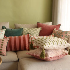 A collection of decorative throw pillows in various colors and patterns, including stripes, checks, and tassels, arranged on a beige sofa against a sage green wall.