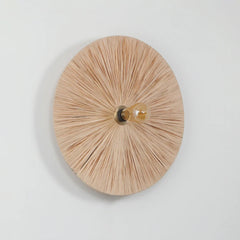 A circular Vagu wall sconce with a natural raffia featuring radiating slats and a single exposed filament light bulb in the center, mounted on a wall.