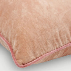 A close-up of a blush pink velvet throw pillow with a matching pink piped edge, shown on a light gray background.