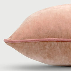 A close-up of a dusty pink velvet throw pillow with a matching pink piped edge, shown against a light gray background.