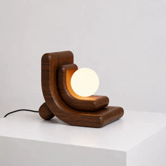 A modern wooden table lamp with a sculptural crescent-shaped dark wood base and a glowing white spherical bulb, shown on a white surface against a light gray background.