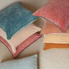 A collection of velvet throw pillows in various colors including teal, dusty pink, peach, orange, and light blue, arranged on a neutral beige textured surface.