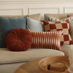 A styled collection of decorative throw pillows on a light beige sofa, featuring a round rust-red pillow, a striped pillow, and a checkered pillow, set against a white paneled wall.