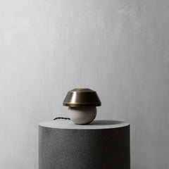 A modern table lamp with a metallic bronze dome-shaped shade and a matte beige spherical base, displayed on a dark gray cylindrical pedestal against a textured light gray wall.