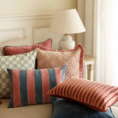 A styled collection of decorative throw pillows in a warm, neutral-toned bedroom setting. The pillows feature various patterns including stripes, checkers, and textured floral motifs in dusty pink, teal blue, rust orange, and cream.