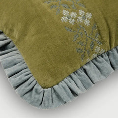 Close-up of a decorative throw pillow in olive green fabric with white floral embroidery and a dusty blue-gray ruffled border, shown on a light gray background.
